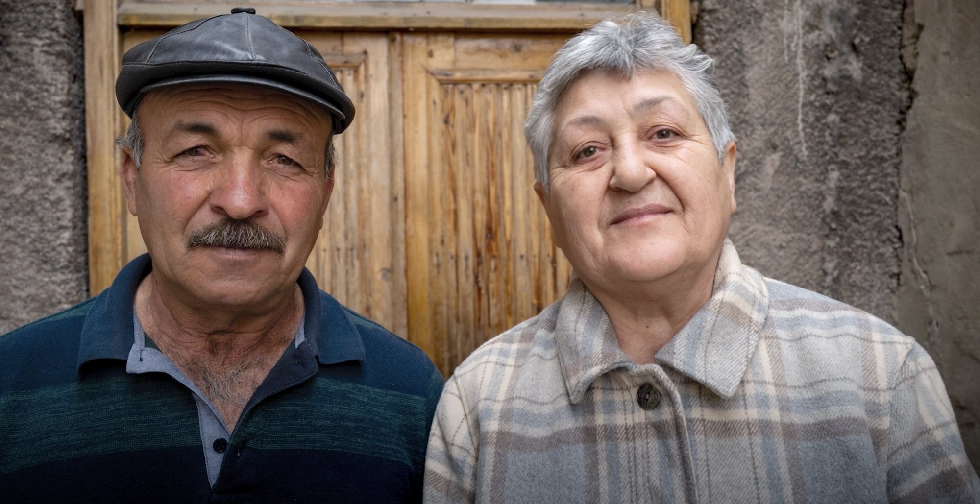 An elderly couple stands in front of a wooden door, looking into the camera