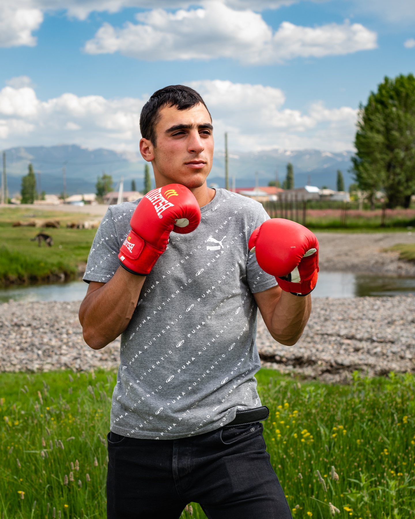 A young man in red boxing gloves stands in a field, mountains behind