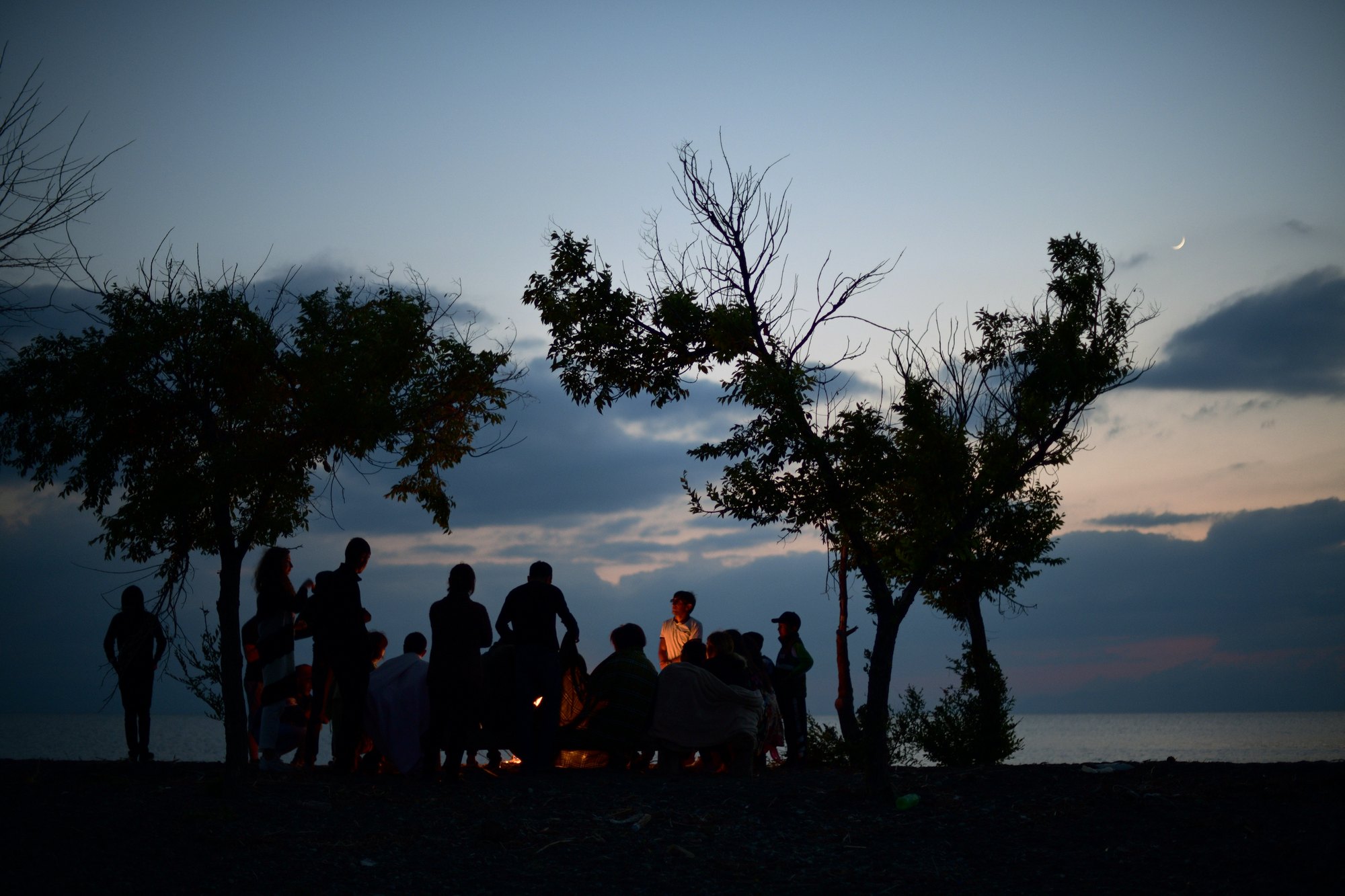 Silhouettes gather round a fire by the water under a crescent moon