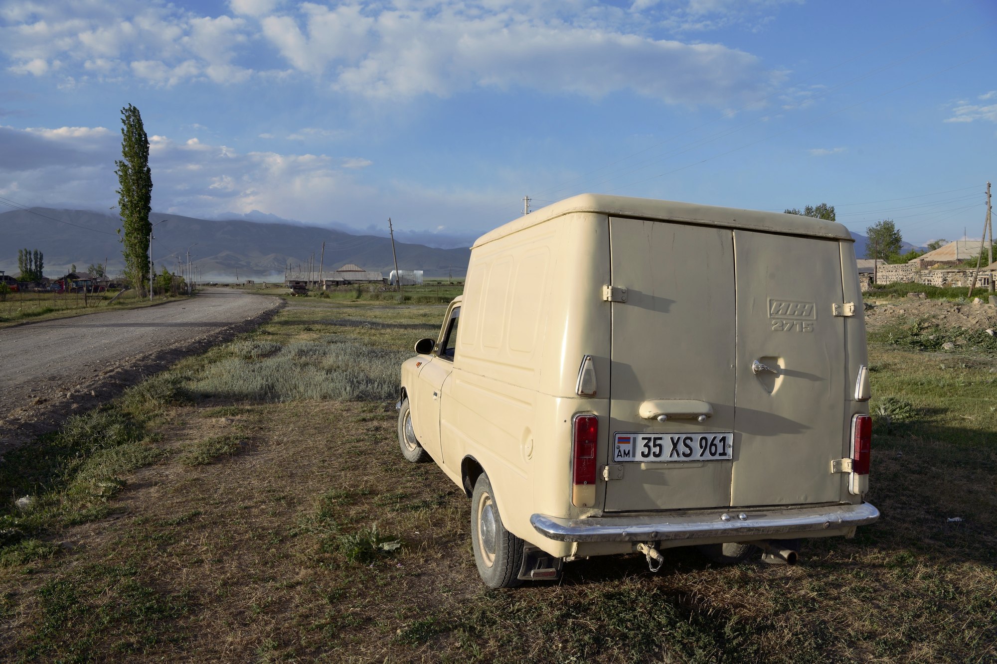 An old cream delivery van parked by a gravel road, mountains behind