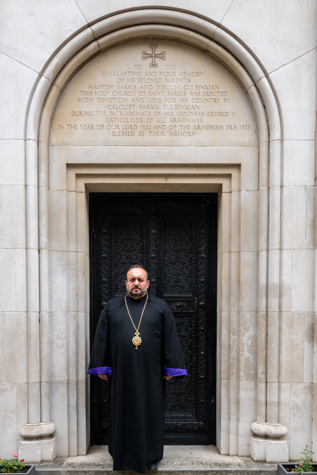 Bishop Hovakim Manukyan in black robes stands in the arched doorway of a pale stone church
