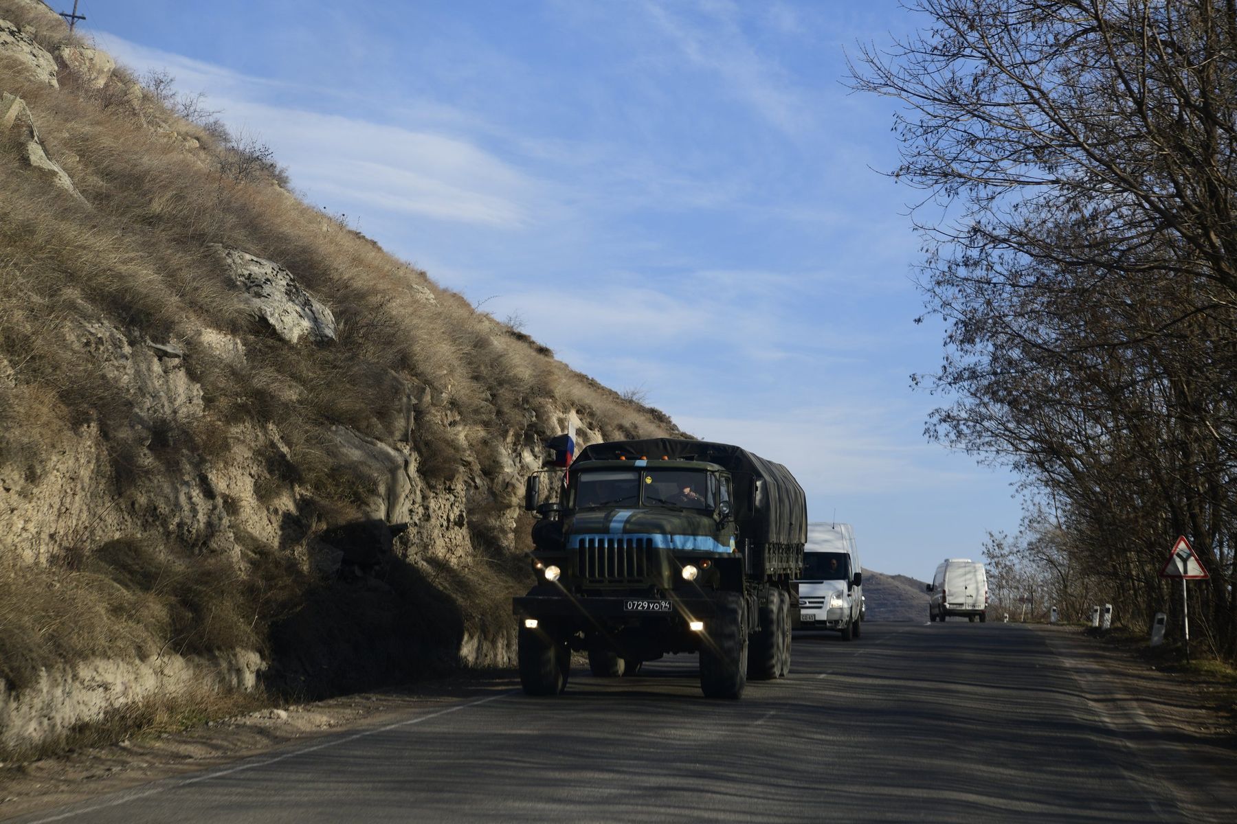 Military trucks drive along a mountain road under a blue sky