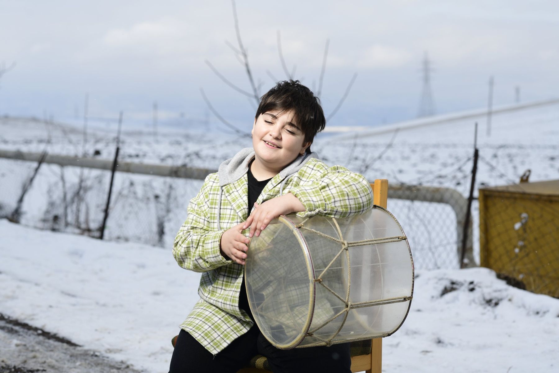 A boy with a frame drum sits on a chair in the snow