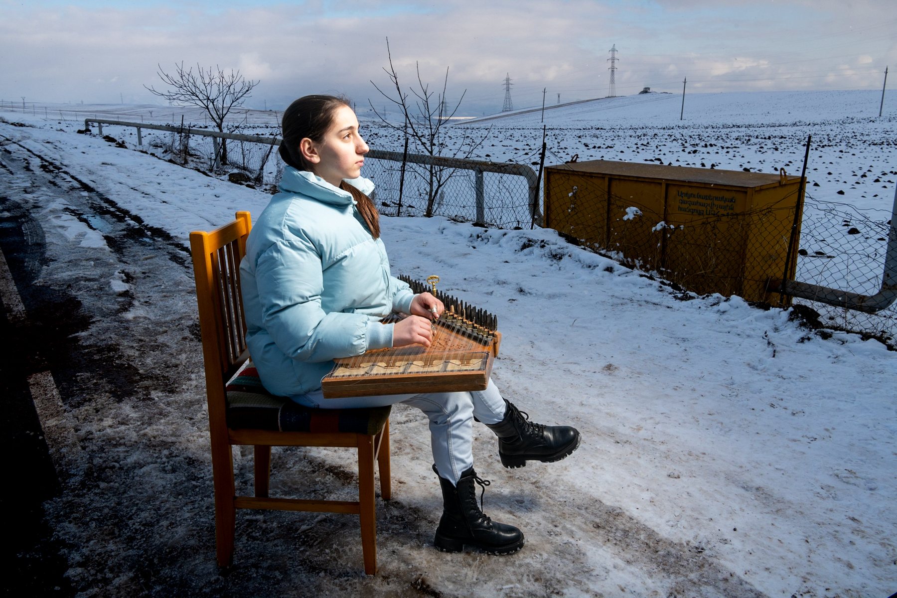 A girl in a pale blue coat plays the qanun on her lap in the snow