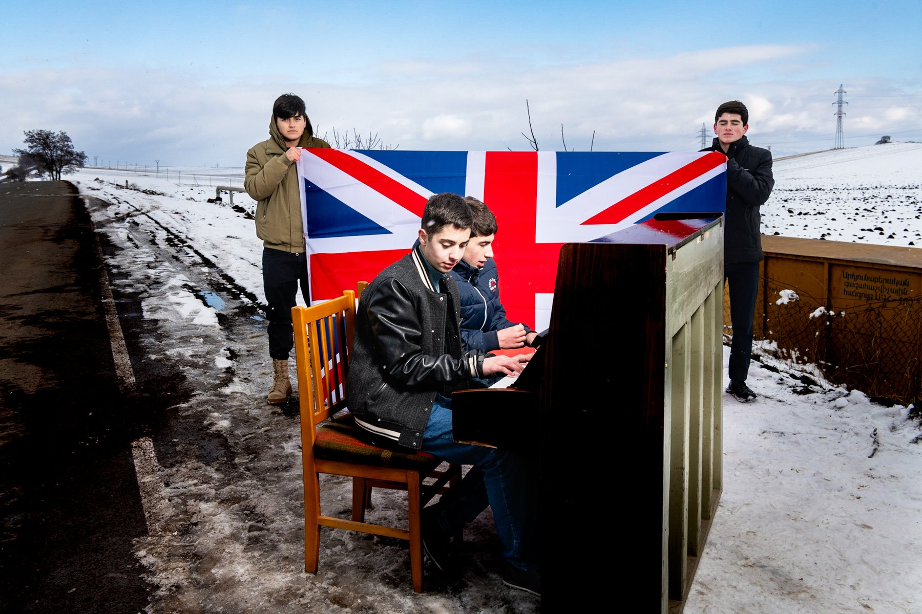 A boy plays an upright piano in the snow, a Union flag held up behind him