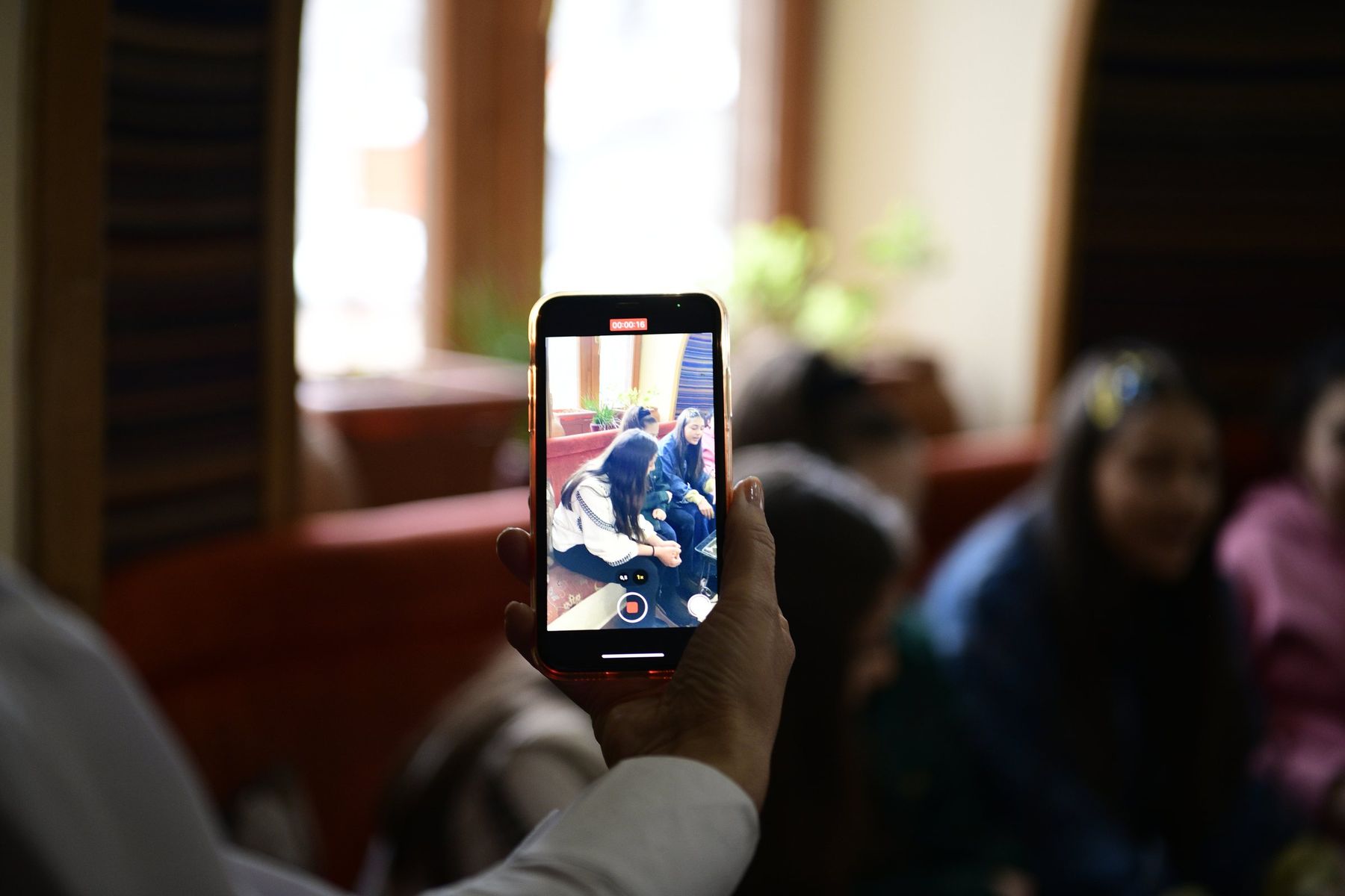 A hand holds up a phone on a video call while girls sit on a sofa