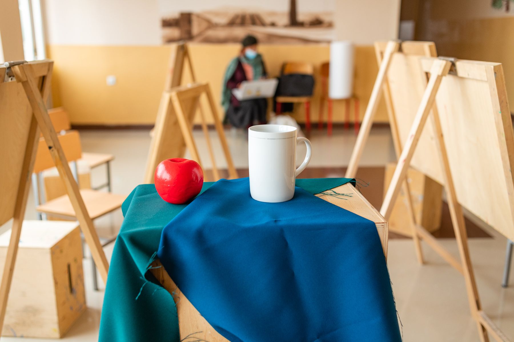 An apple and a white jug on a blue cloth, easels behind
