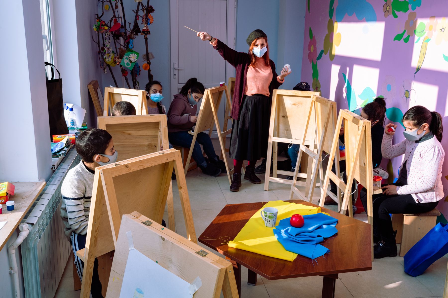 A teacher points across a room of children at easels