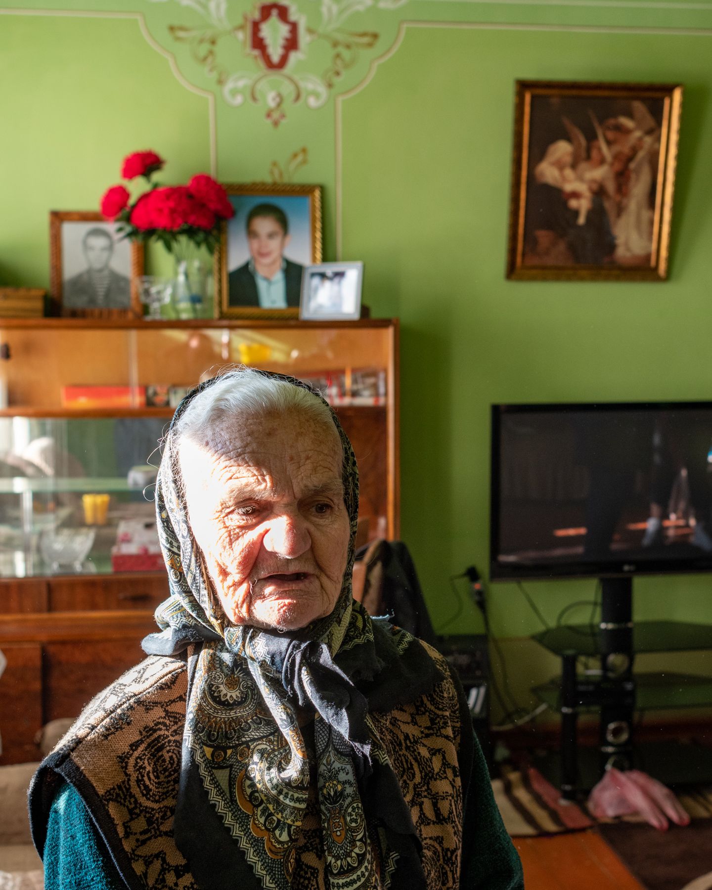 An elderly woman in a headscarf beneath framed portraits
