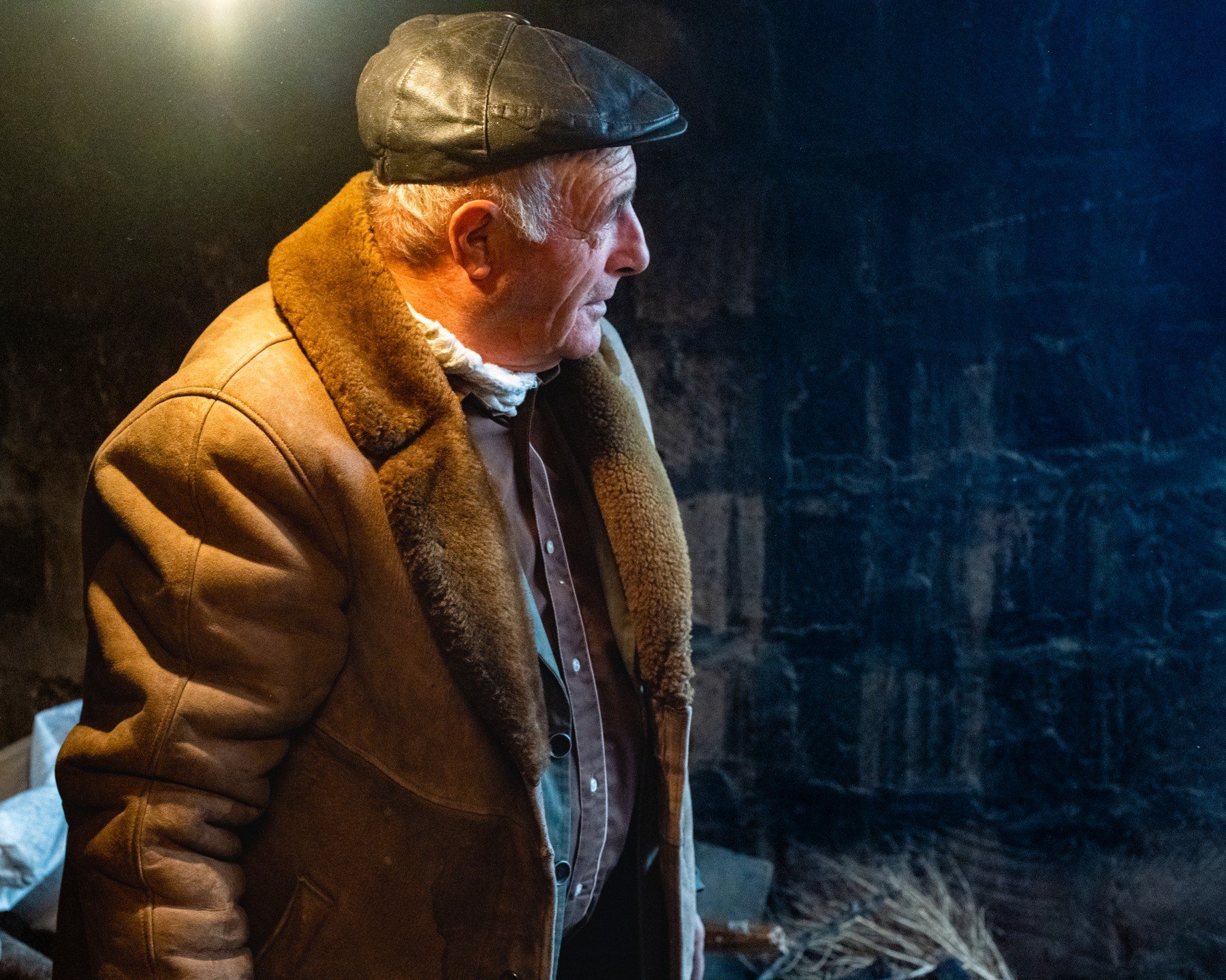 An older man in a leather cap and sheepskin coat stands in a dark stone room