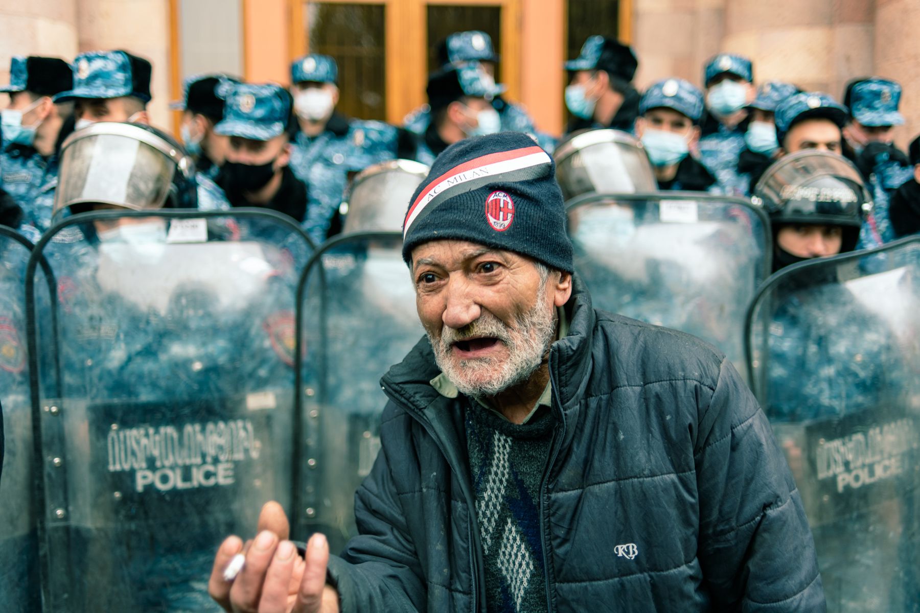 An older man gestures in front of police riot shields