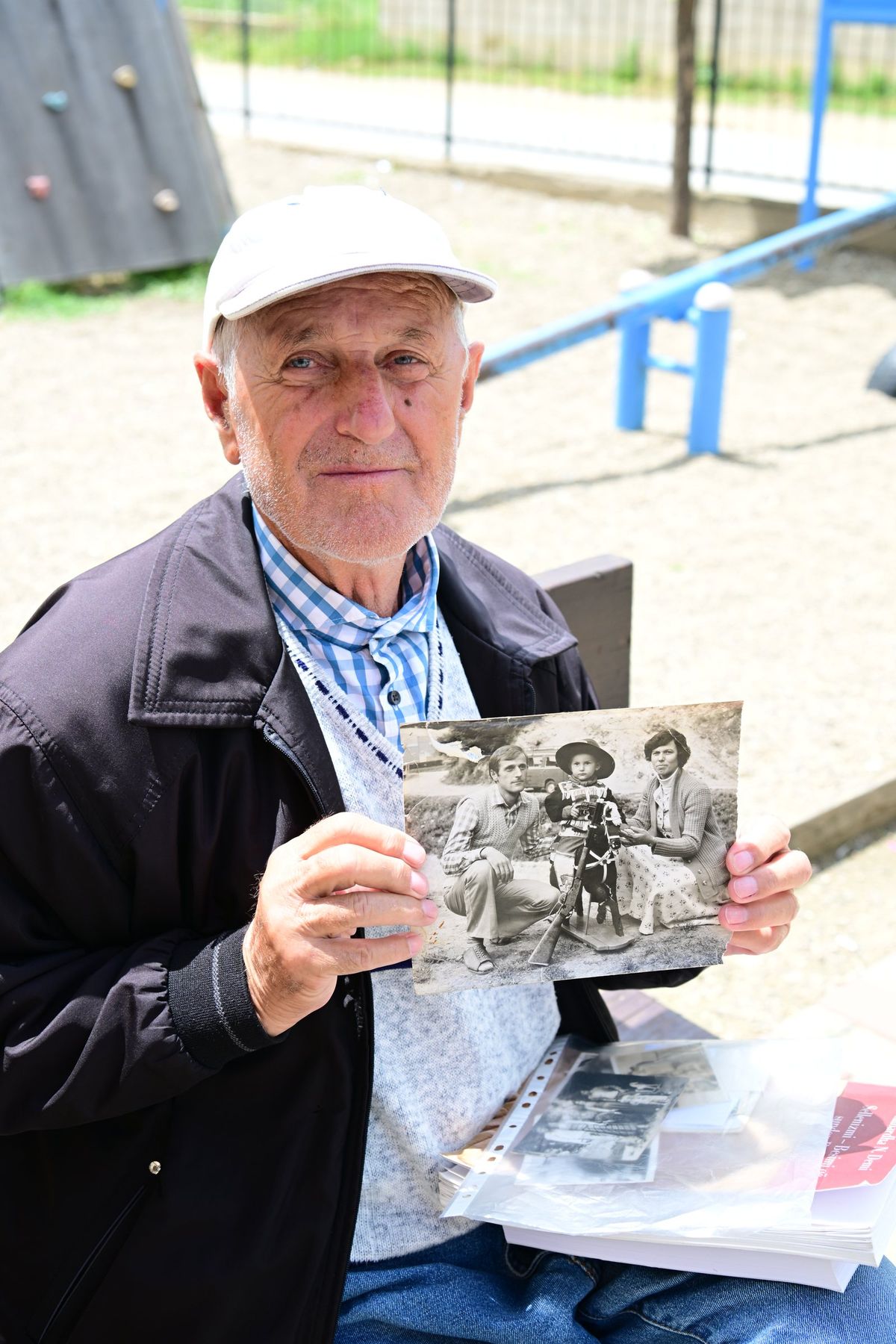 An elderly man in a white cap holds up an old black and white photograph