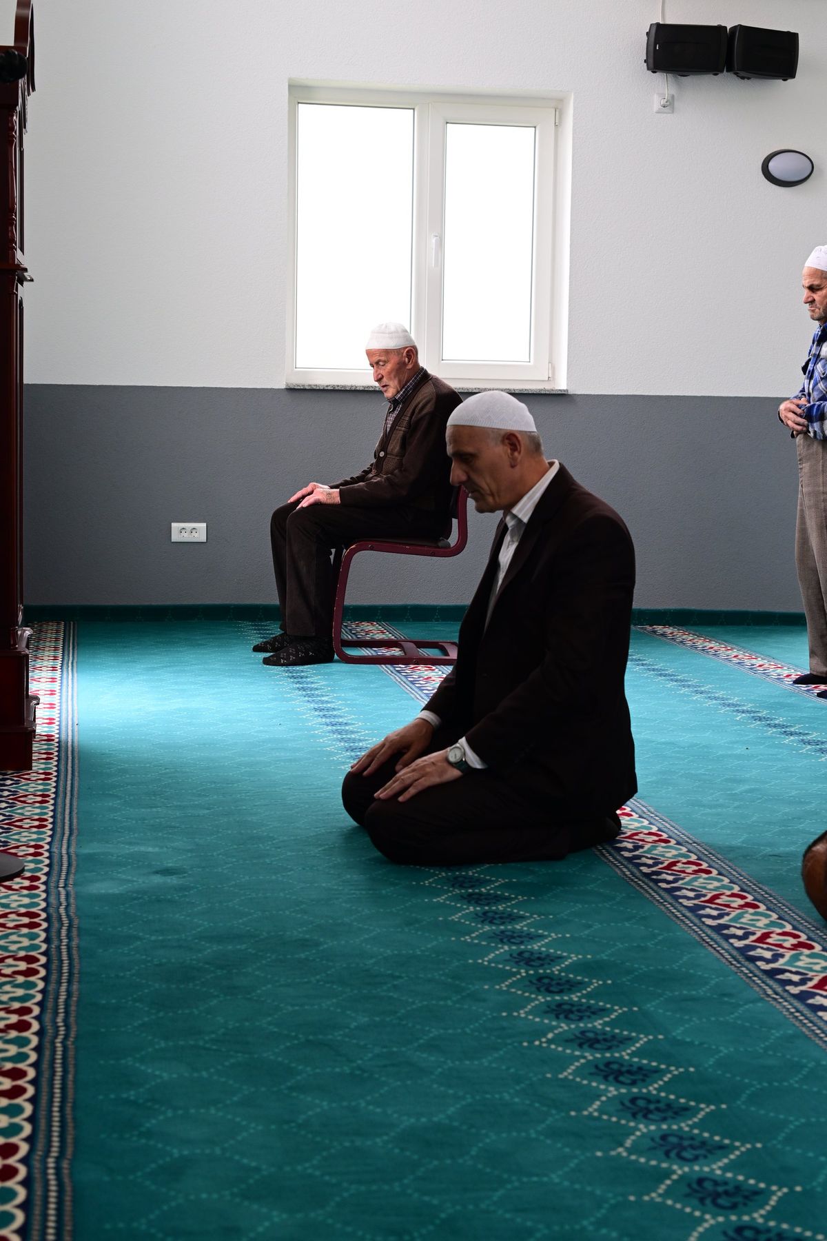 A man kneels on a turquoise carpet in a bright mosque