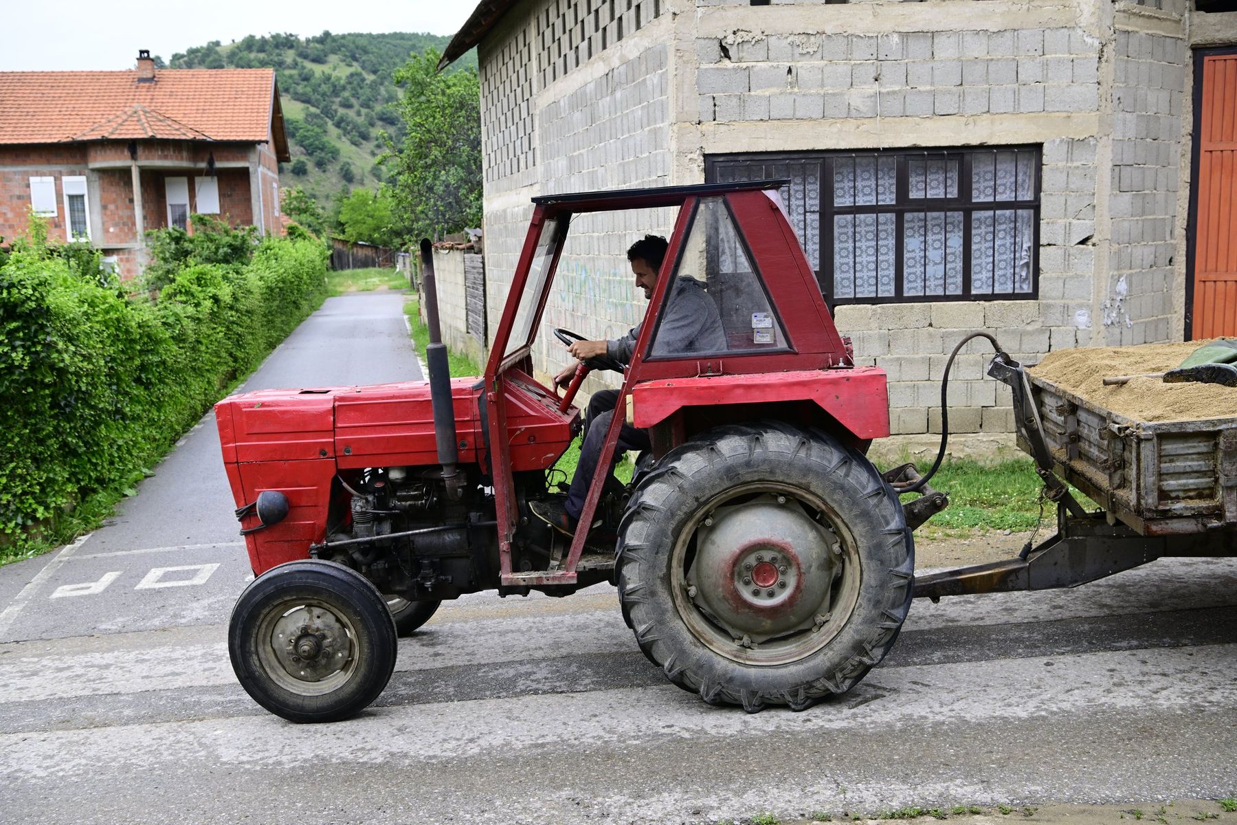 A man drives a red tractor and trailer down a village lane