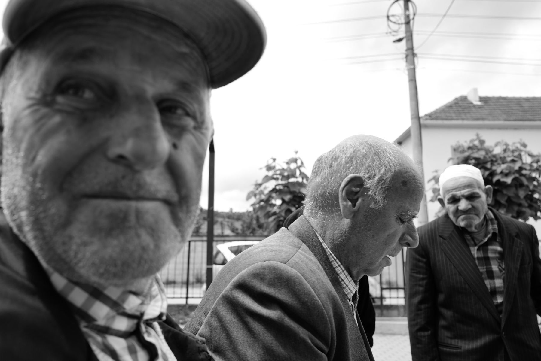 Black and white: an old man smiles close to the camera, two others talk behind him