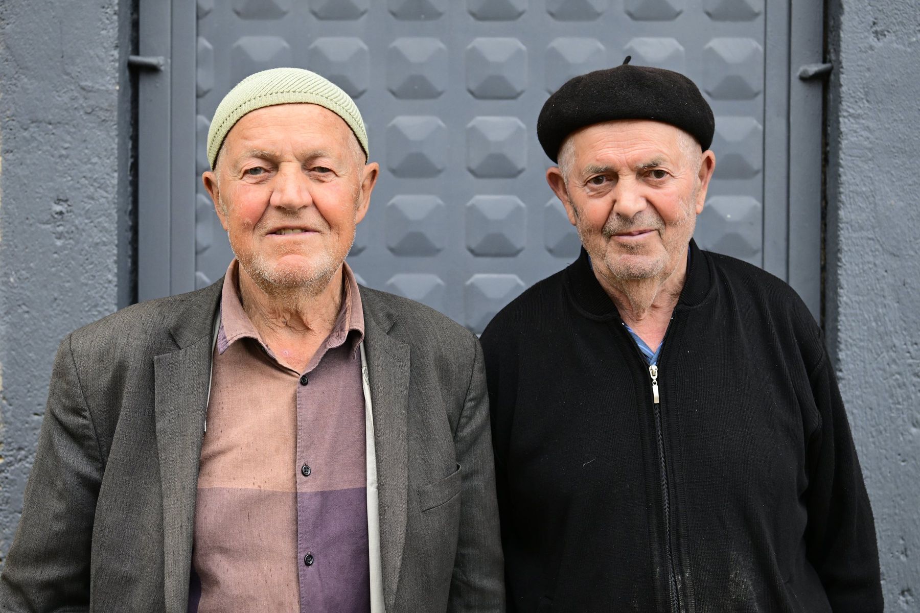 Two elderly men in caps stand side by side before a metal gate