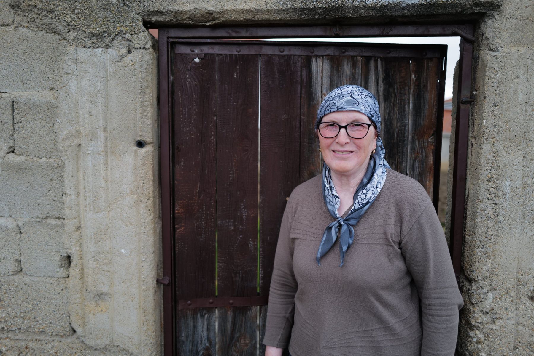 A woman in a headscarf stands smiling before a wooden door