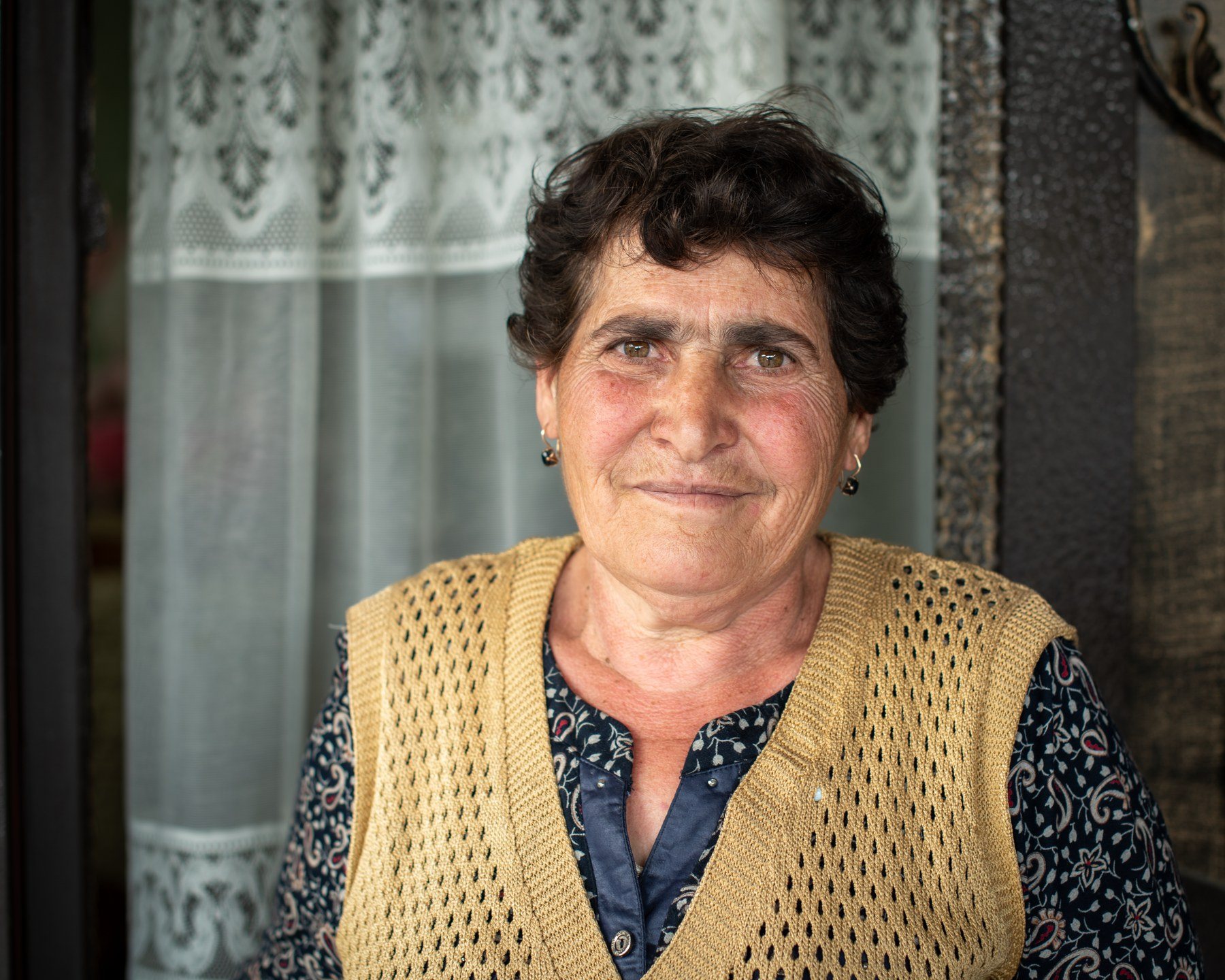 A woman in a knitted waistcoat stands in a doorway, a lace curtain behind her
