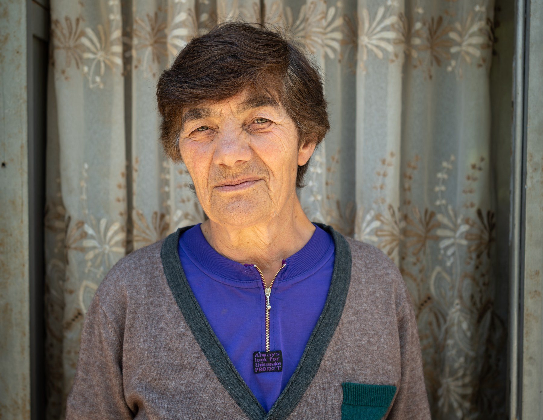 An older woman in a grey cardigan stands before a floral curtain