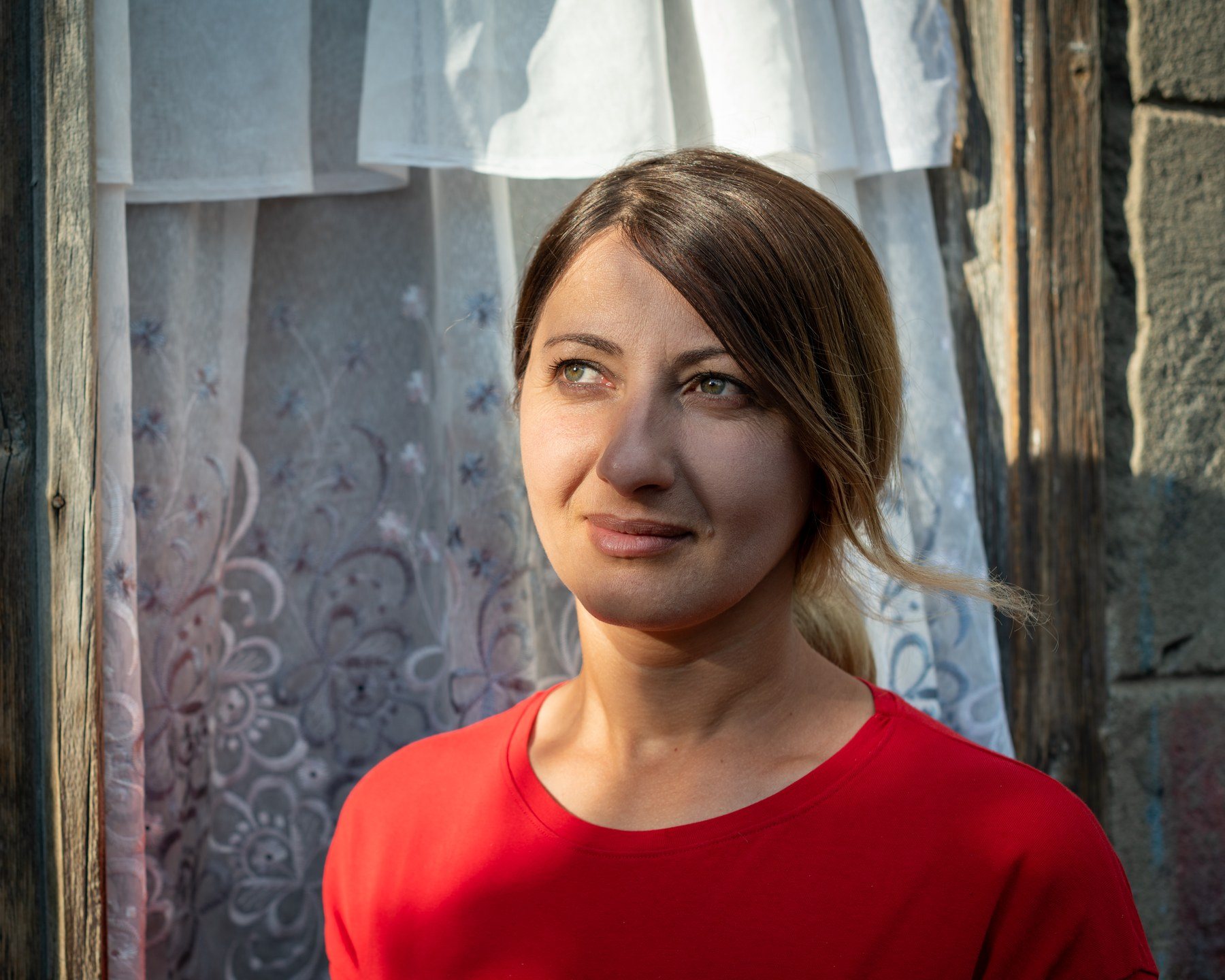 A young woman in a red top looks past the camera, white curtains behind