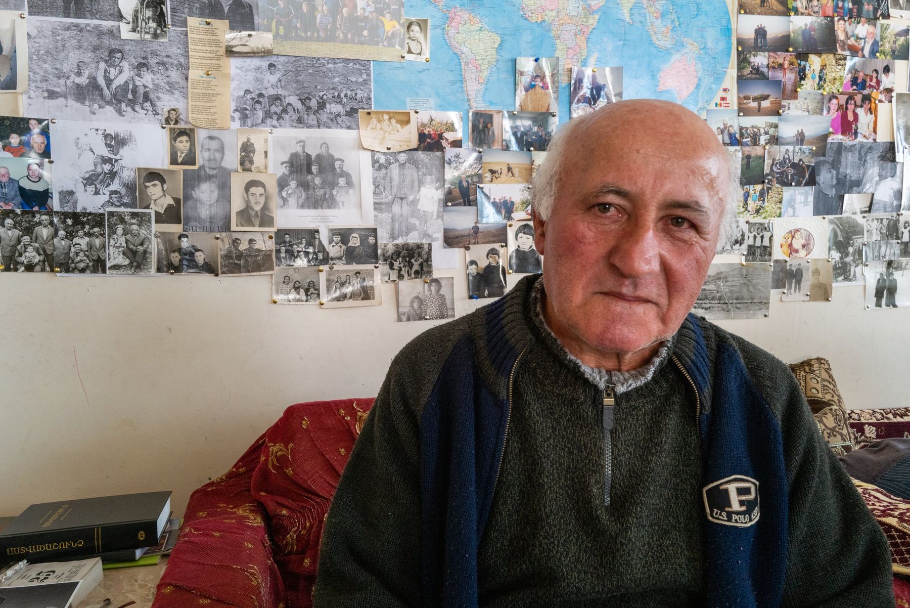An elderly man sits in front of a wall covered in old family and village photographs