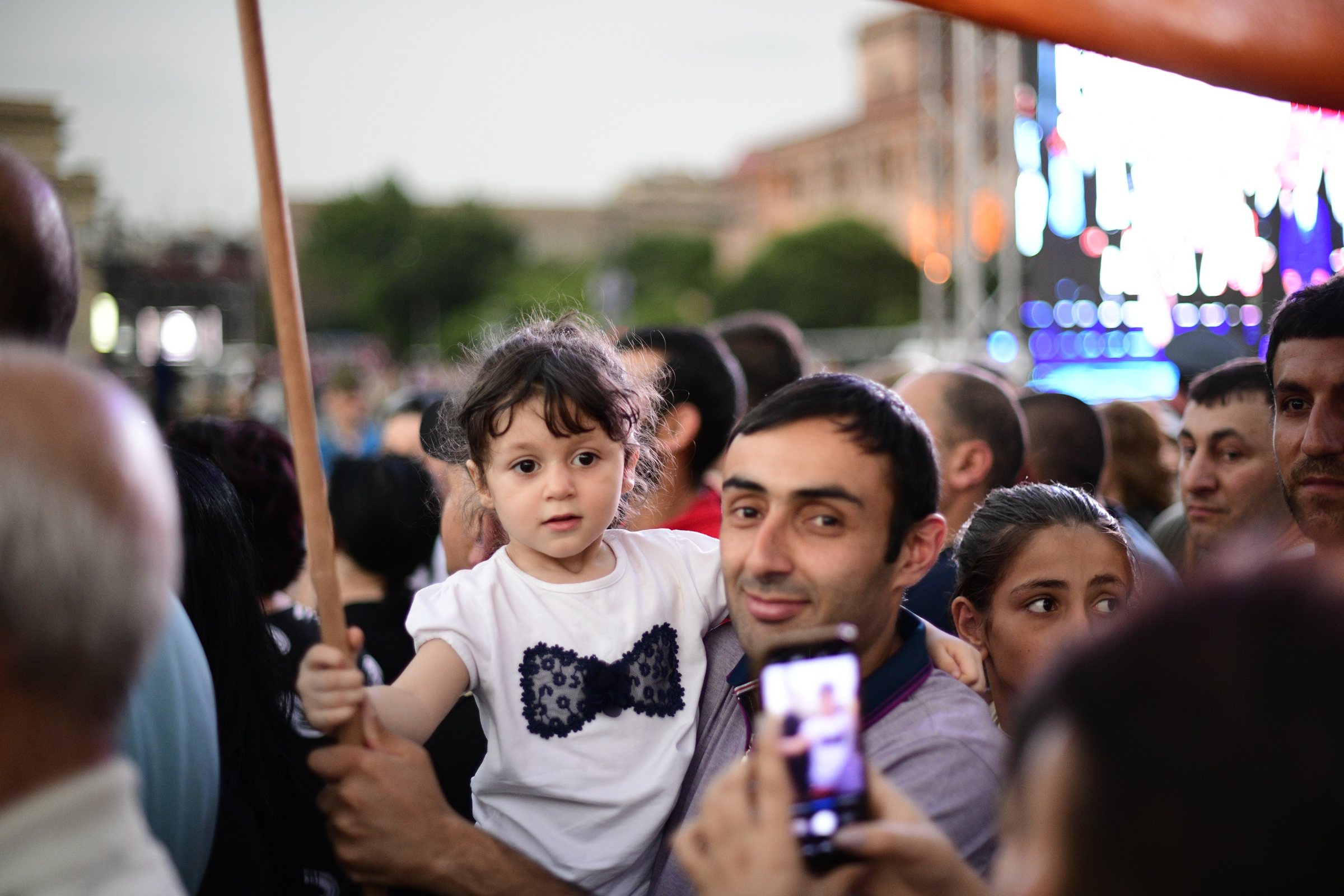 A small girl in a bow-tie T-shirt is held up in the crowd beside her father
