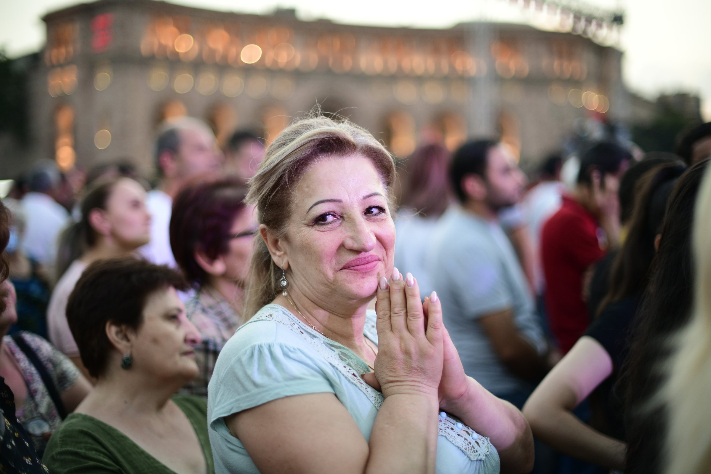 A woman in the crowd presses her hands together and smiles at the camera, the lit government buildings behind