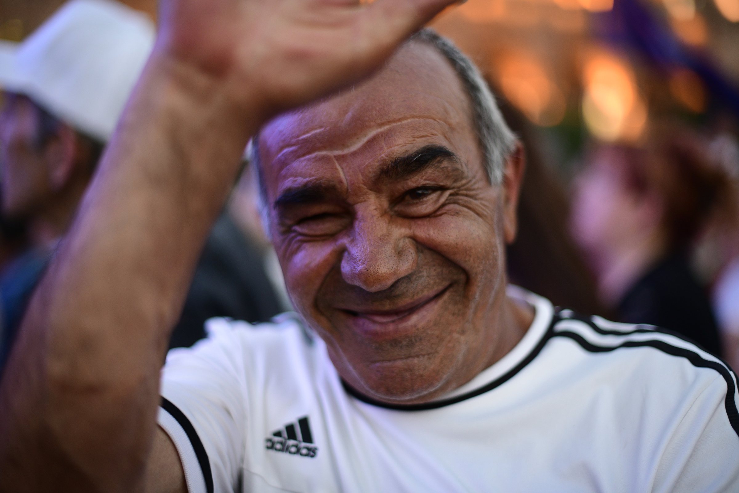 An older man in a white sports shirt waves and laughs into the camera
