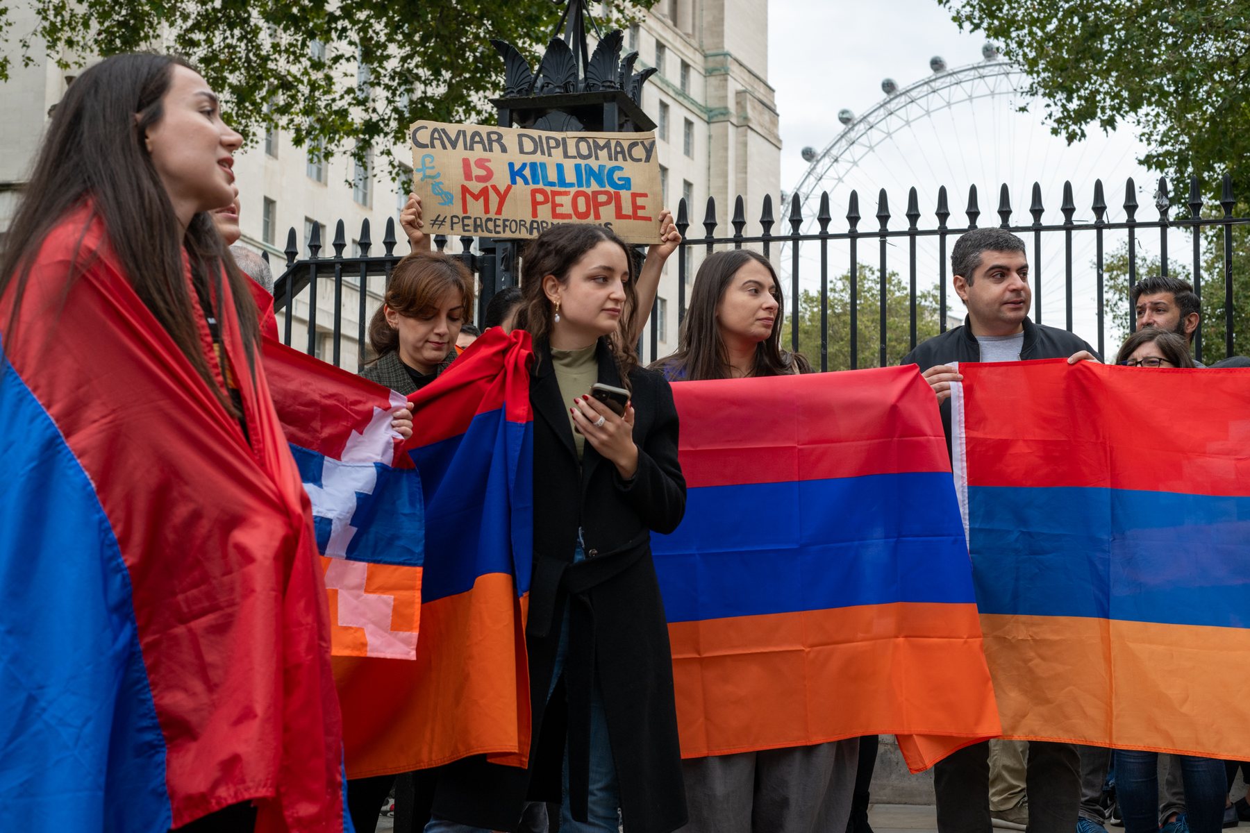 A young woman wrapped in the Artsakh flag stands at the railings among other protesters
