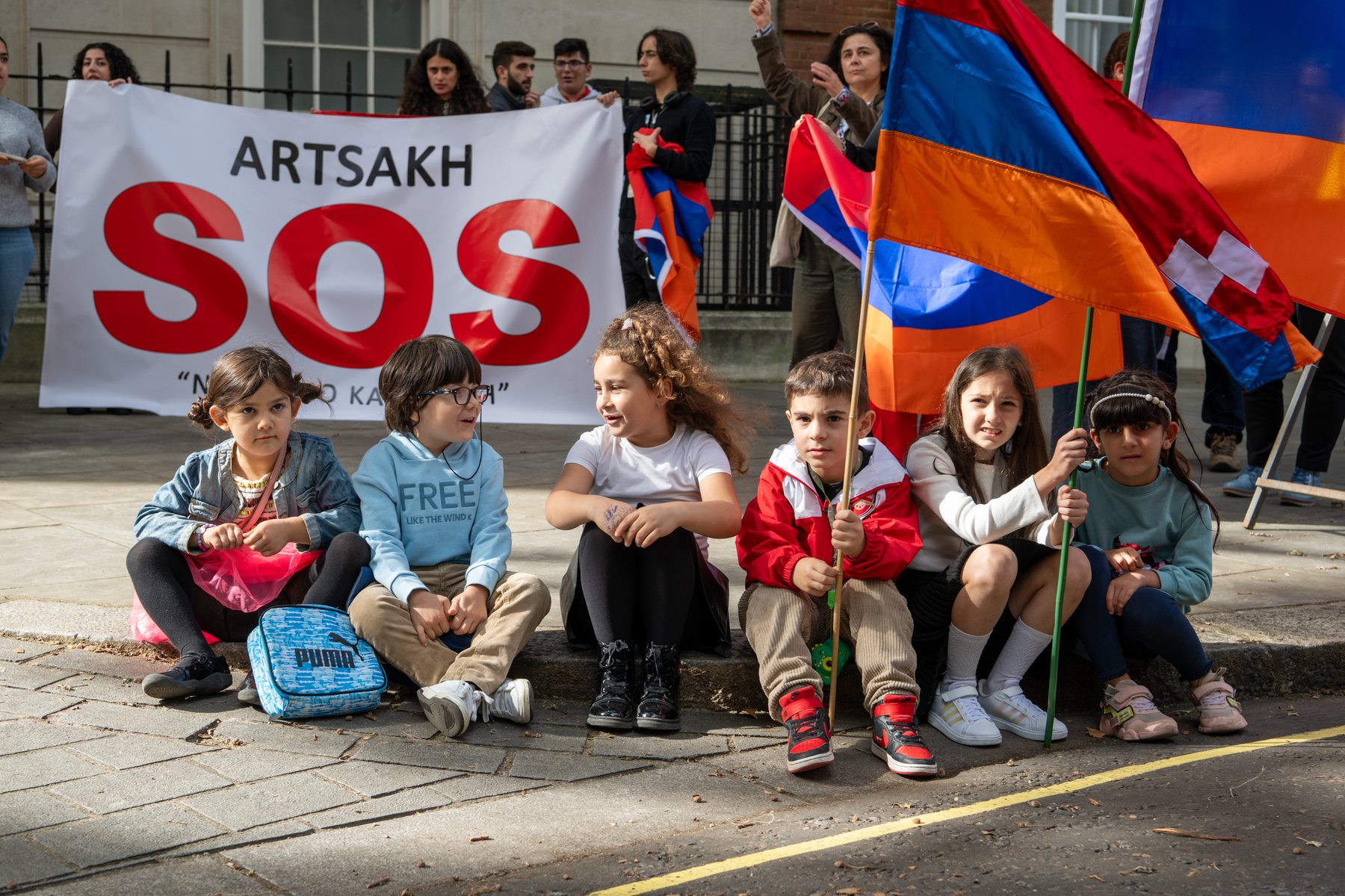 Children sit on the pavement beneath an Artsakh SOS banner