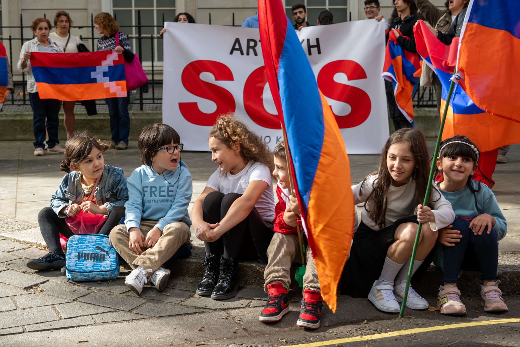 Children sit on the pavement beside an Artsakh SOS banner and flags