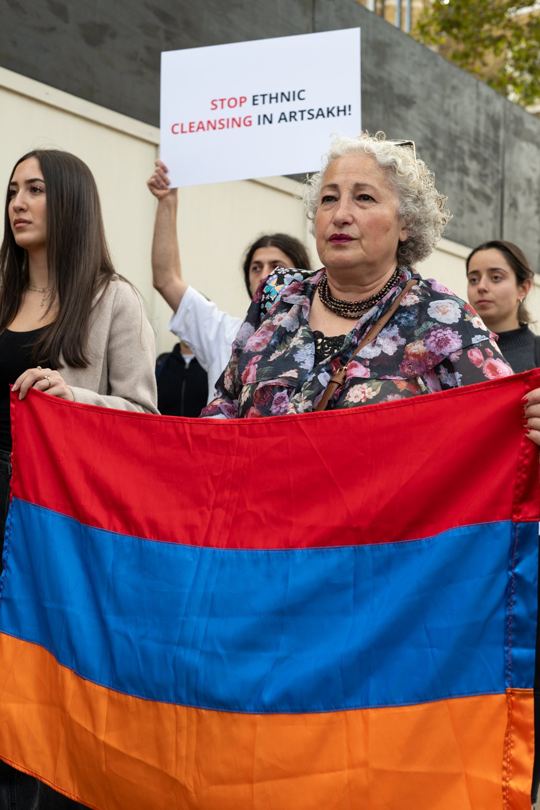 An older woman holds the Armenian flag beneath a sign that reads Stop ethnic cleansing in Artsakh