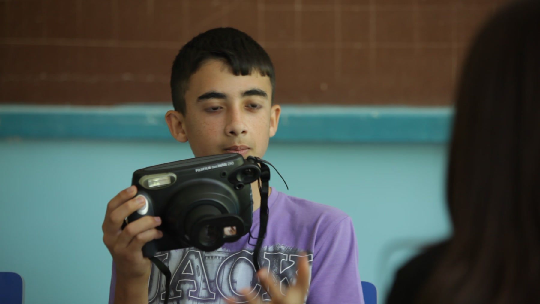 A boy in a purple T-shirt holds the Instax camera up in both hands