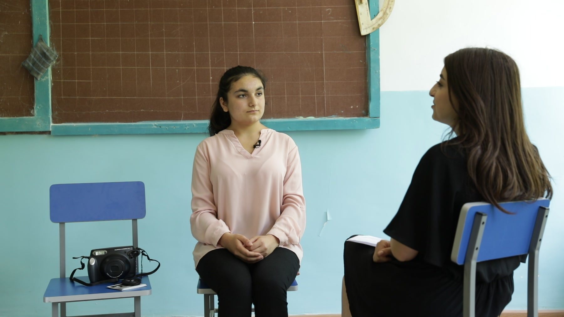 A girl sits on a chair in a classroom, talking with a young woman, an Instax camera on the chair beside her