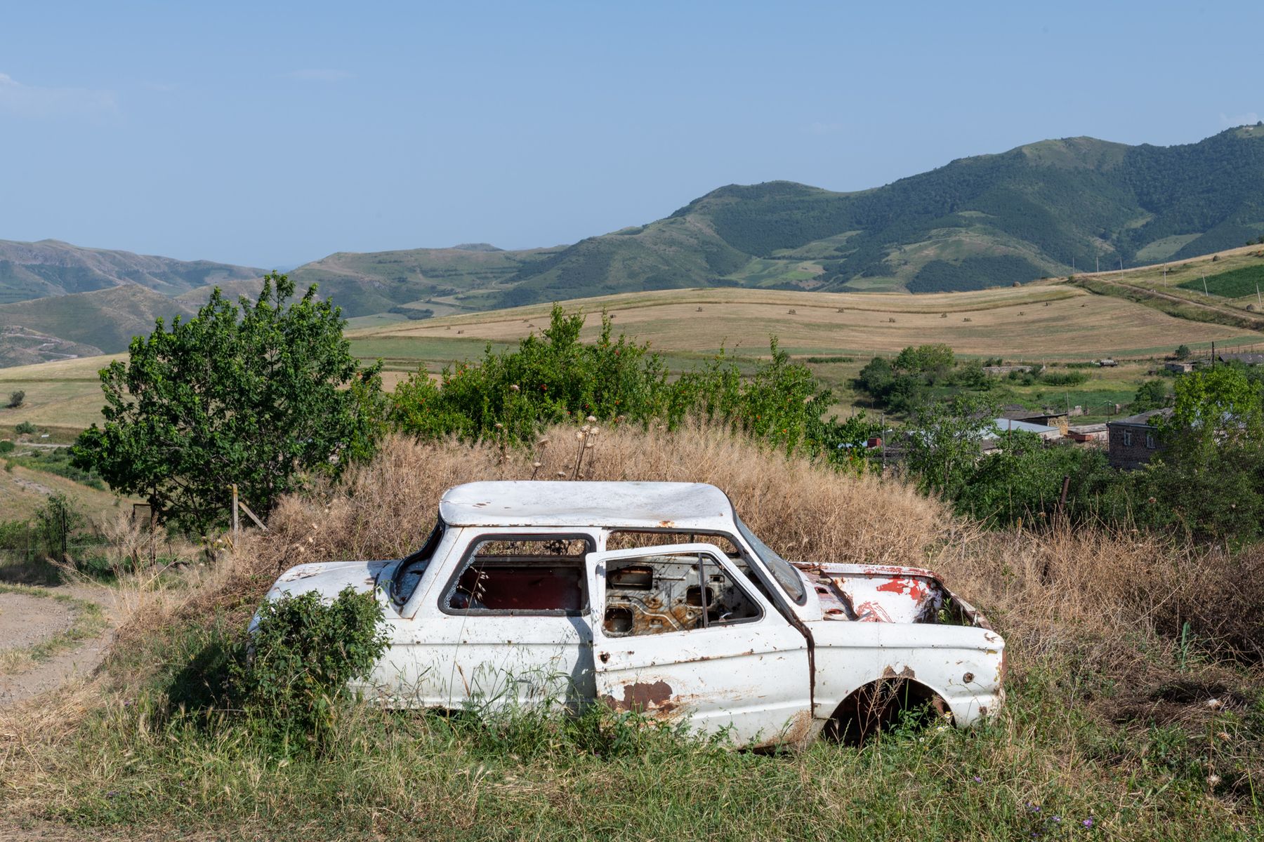 A wrecked white car in long grass, hills behind