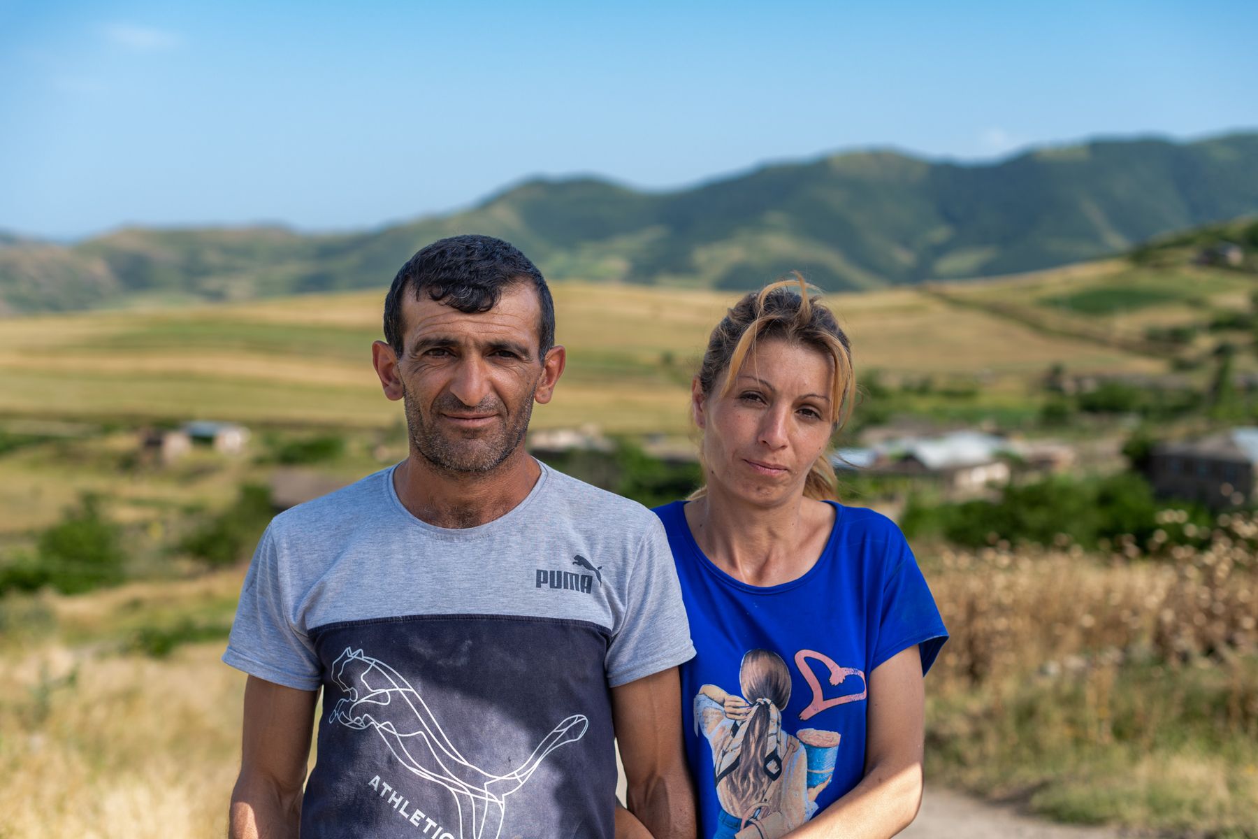 A man and a woman stand side by side, green hills behind