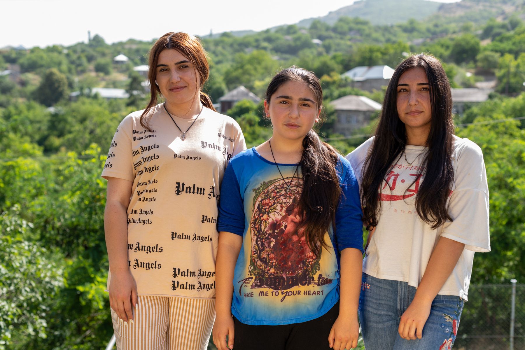 Three teenage girls stand among trees above the village